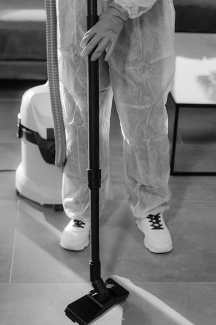 A person wearing protective clothing, including gloves and a disposable suit, is using a vacuum cleaner with a long wand attachment to perform surface cleaning on a smooth, hard floor. The scene is se