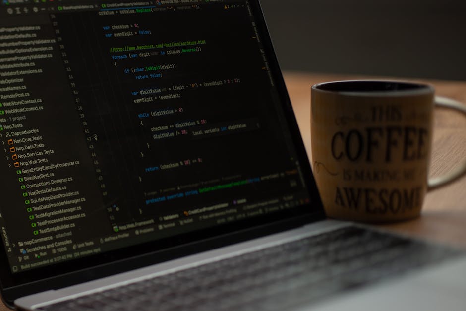 A black and white image depicting a workspace with a laptop open to a screen displaying lines of code, placed on a dark wooden desk against a textured brick wall background. To the right of the laptop