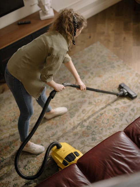A woman performing surface cleaning with a yellow canister vacuum cleaner on an ornate, patterned area rug in a living room. The rug features a floral design with shades of beige, green, and muted ora